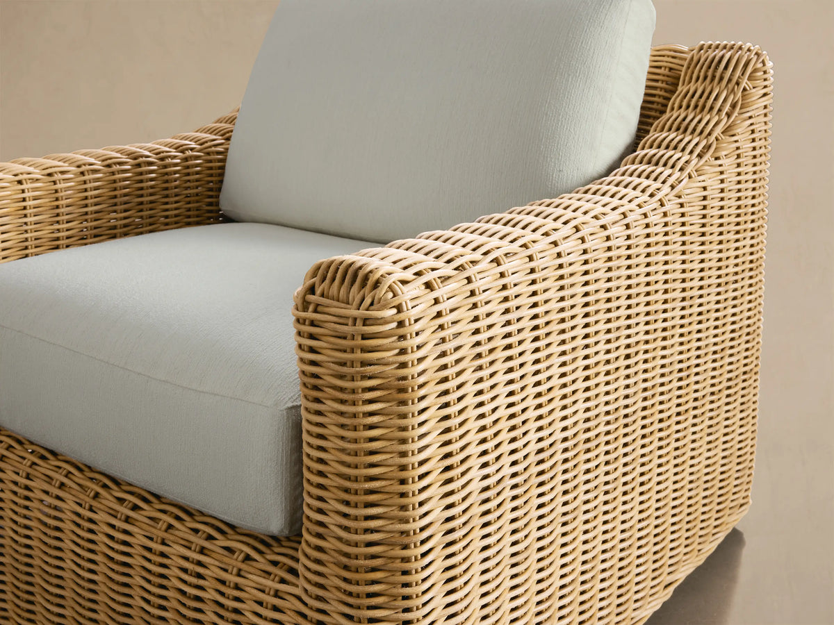 Montauk Outdoor Swivel Chair with natural woven wicker base & arms, light cream seat & back cushions, on a patio.