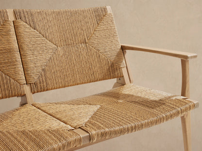 Mustique Outdoor Sofa, light golden-toned teak wood frame, natural woven wicker seat/back with diamond pattern, on stone patio.