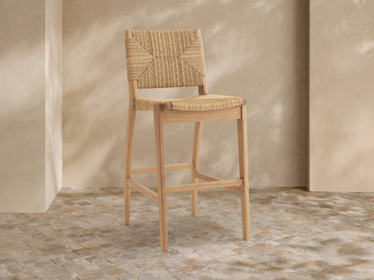 Mustique Outdoor Barstool with natural teak frame, woven tan seat & backrest, angled left on a light tiled floor.