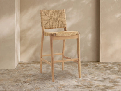 Mustique Outdoor Barstool with natural teak frame, woven tan seat & backrest, angled left on a light tiled floor.