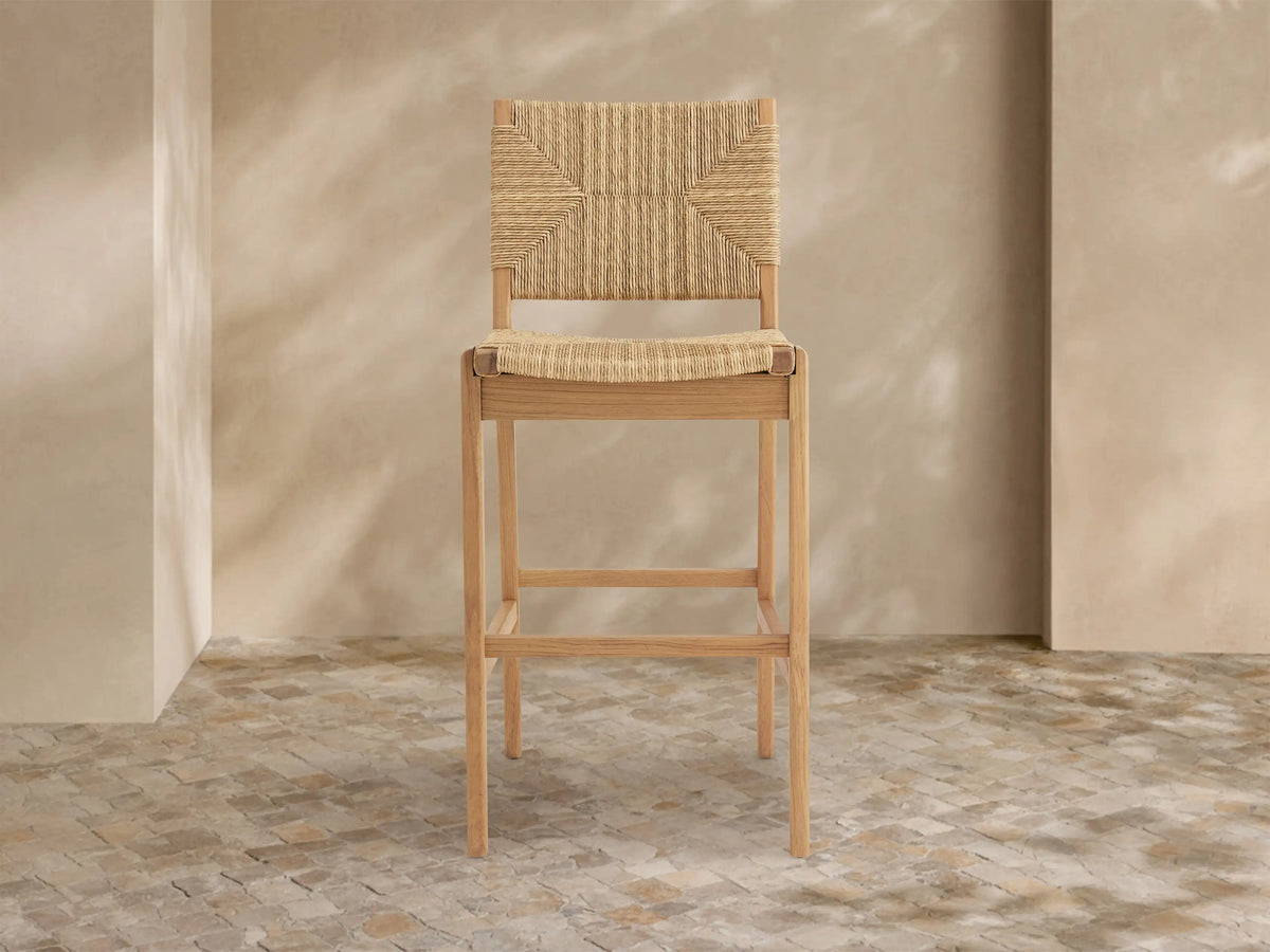 Mustique Outdoor Barstool with natural teak frame, woven tan seat & backrest, angled left on a light tiled floor.