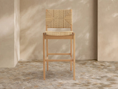 Mustique Outdoor Barstool with natural teak frame, woven tan seat & backrest, angled left on a light tiled floor.