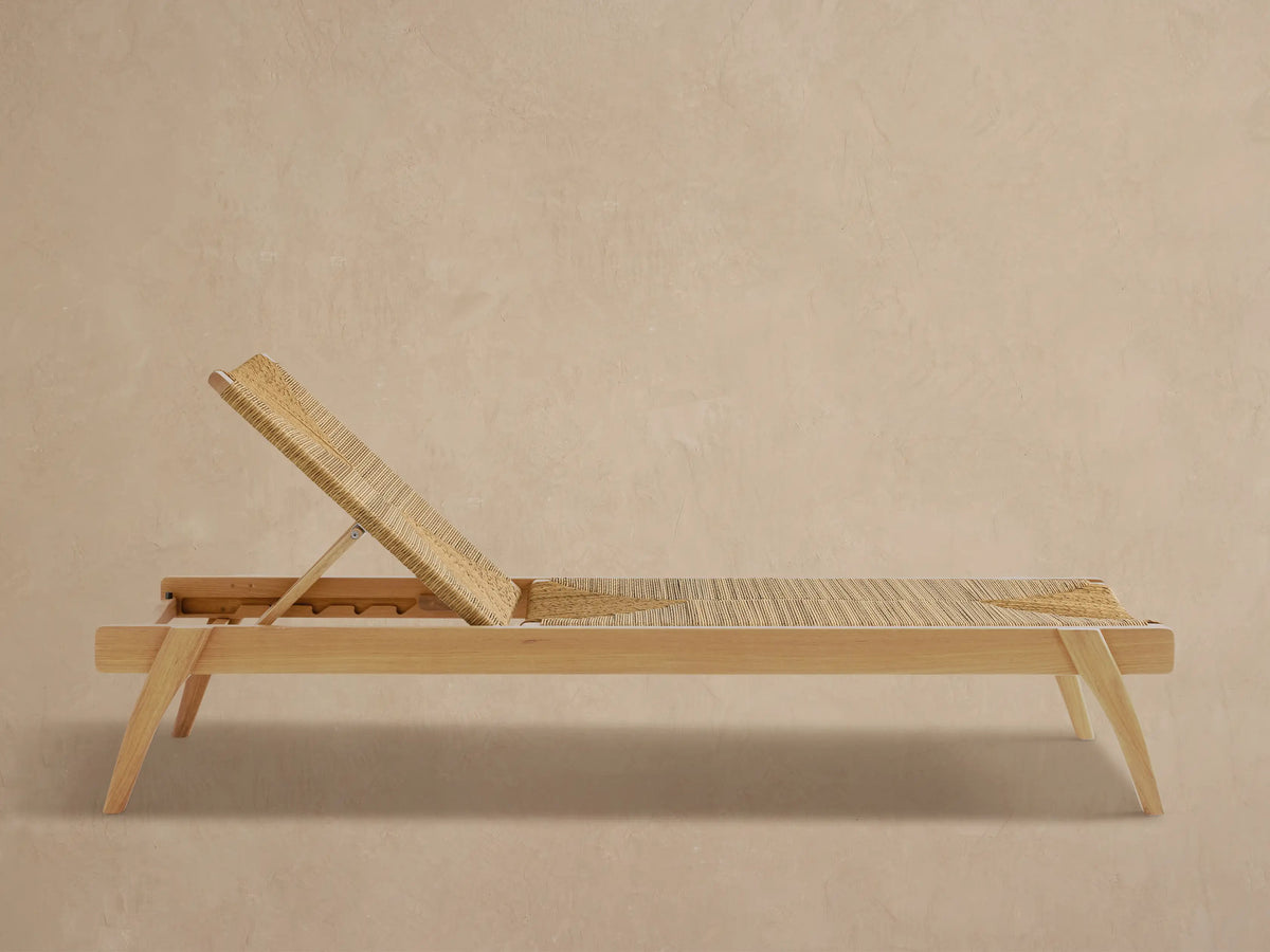 Mustique Outdoor Chaise, light teak frame, natural woven seat & angled backrest with geometric pattern, on paved patio.