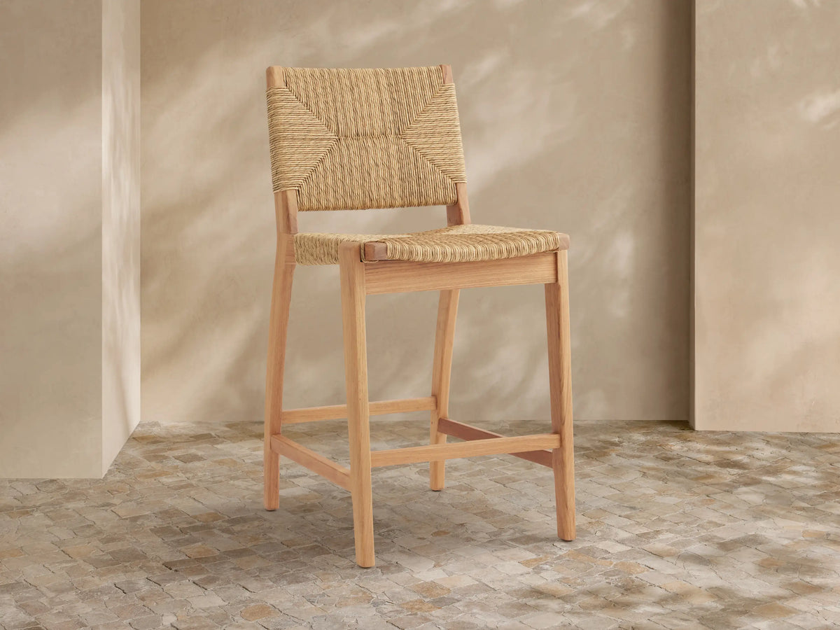 Light teak Mustique Outdoor Counter Stool with woven beige wicker back & seat, angled left against a light wall on stone tiles.