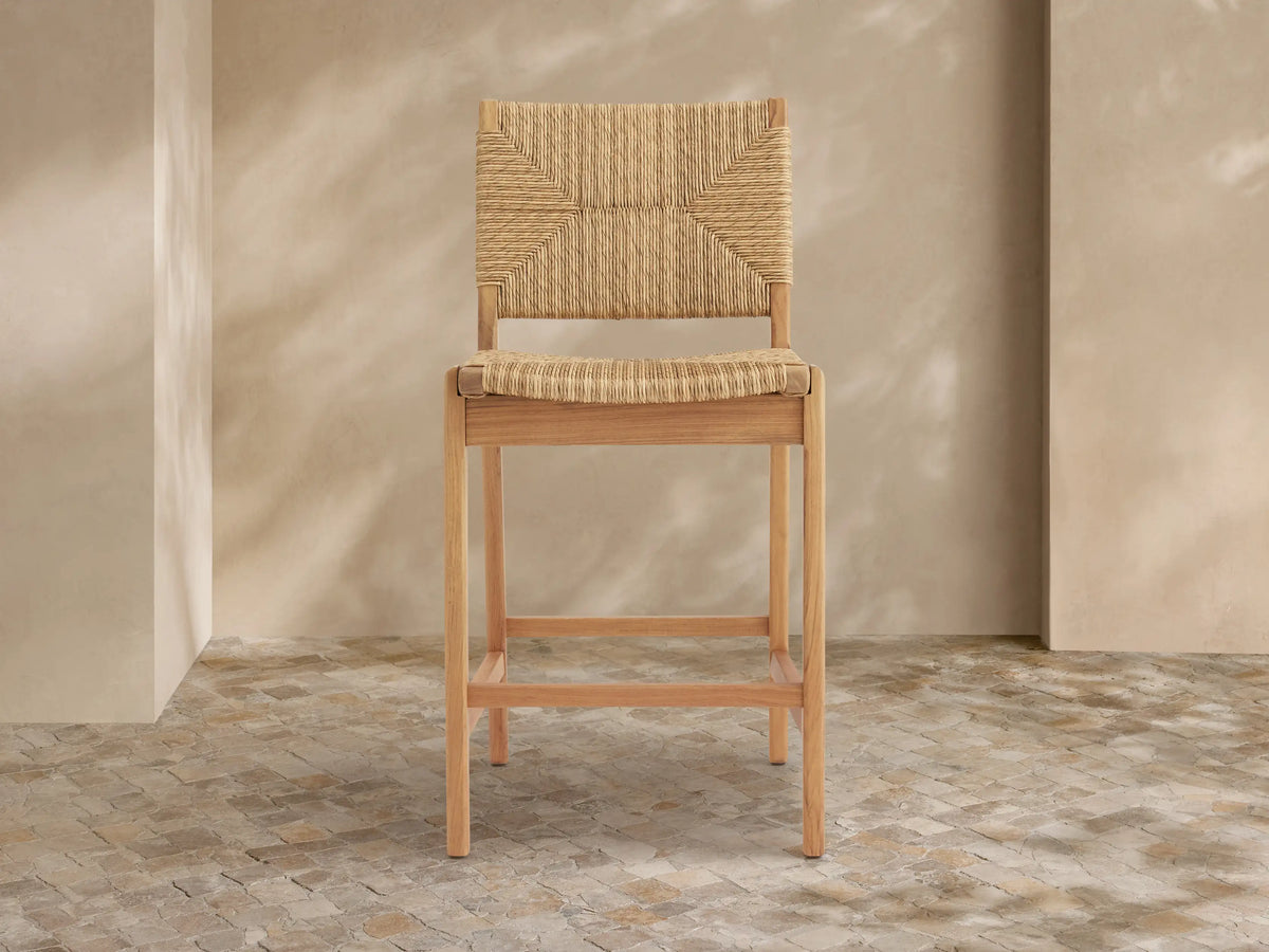 Light teak Mustique Outdoor Counter Stool with woven beige wicker back & seat, angled left against a light wall on stone tiles.
