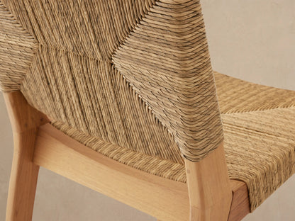Mustique Outdoor Counter Stool, front view, golden teak frame, handwoven natural wicker seat/back, on patterned tile floor.