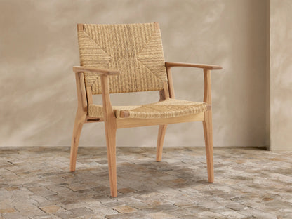 Mustique teak wood outdoor dining arm chair with woven wicker back and seat, angled slightly on a textured tile floor.