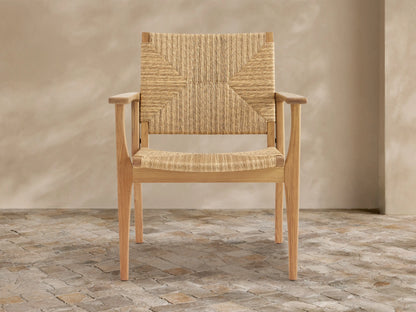 Mustique teak wood outdoor dining arm chair with woven wicker back and seat, angled slightly on a textured tile floor.