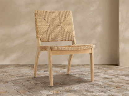 Mustique Outdoor Dining Side Chair with a light teak wood frame, natural tan woven wicker seat & back, angled left on stone floor.