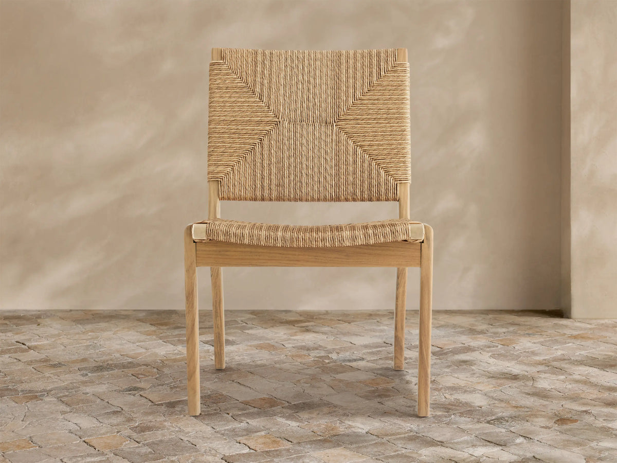 Mustique Outdoor Dining Side Chair with a light teak wood frame, natural tan woven wicker seat & back, angled left on stone floor.