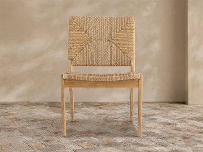 Mustique Outdoor Dining Side Chair with a light teak wood frame, natural tan woven wicker seat & back, angled left on stone floor.