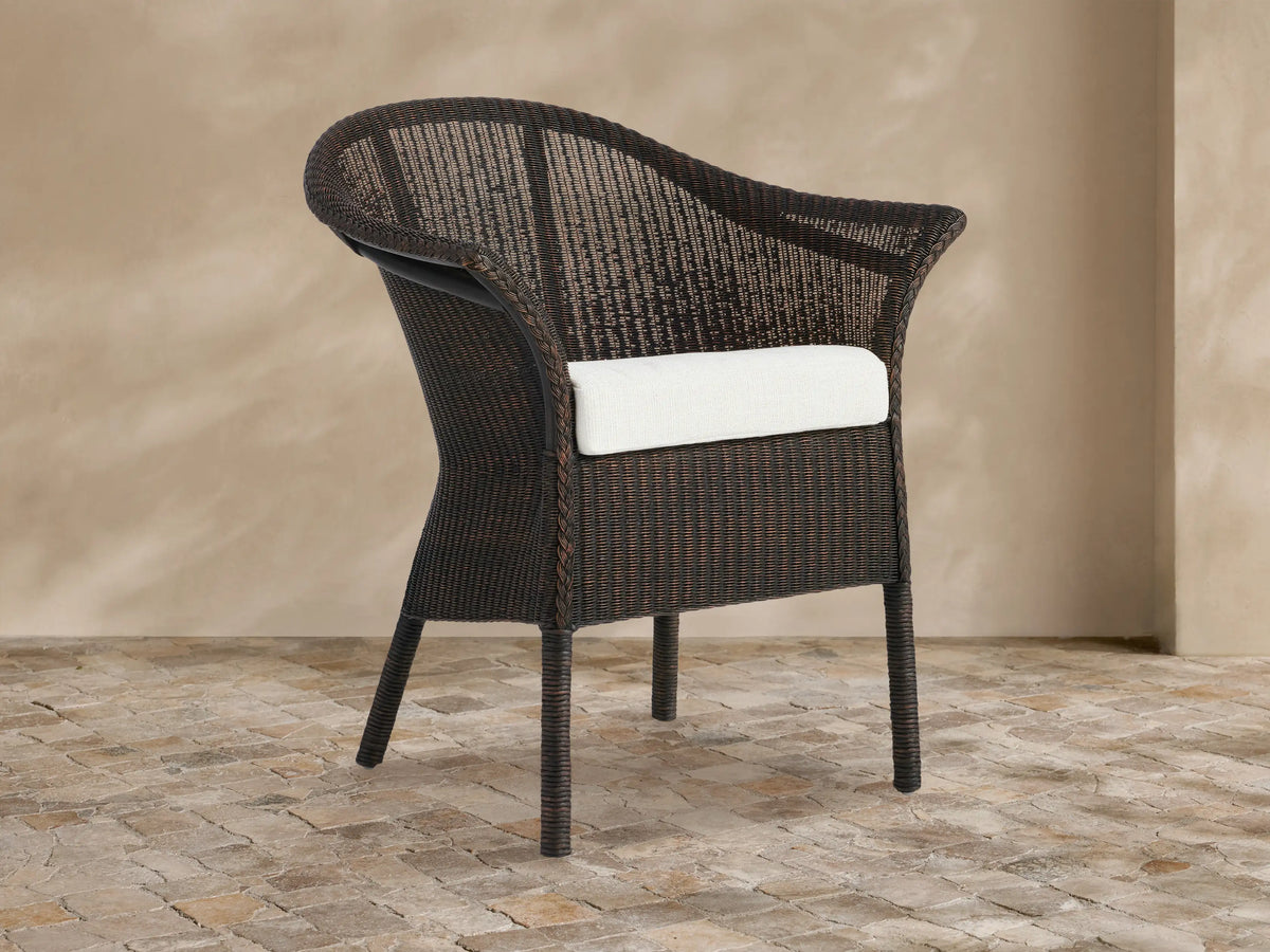 Dark brown woven rattan Nantucket Sunroom Dining Arm Chair with a white seat cushion, angled slightly on a stone floor.