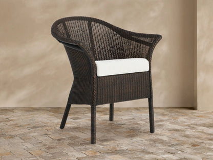 Dark brown woven rattan Nantucket Sunroom Dining Arm Chair with a white seat cushion, angled slightly on a stone floor.