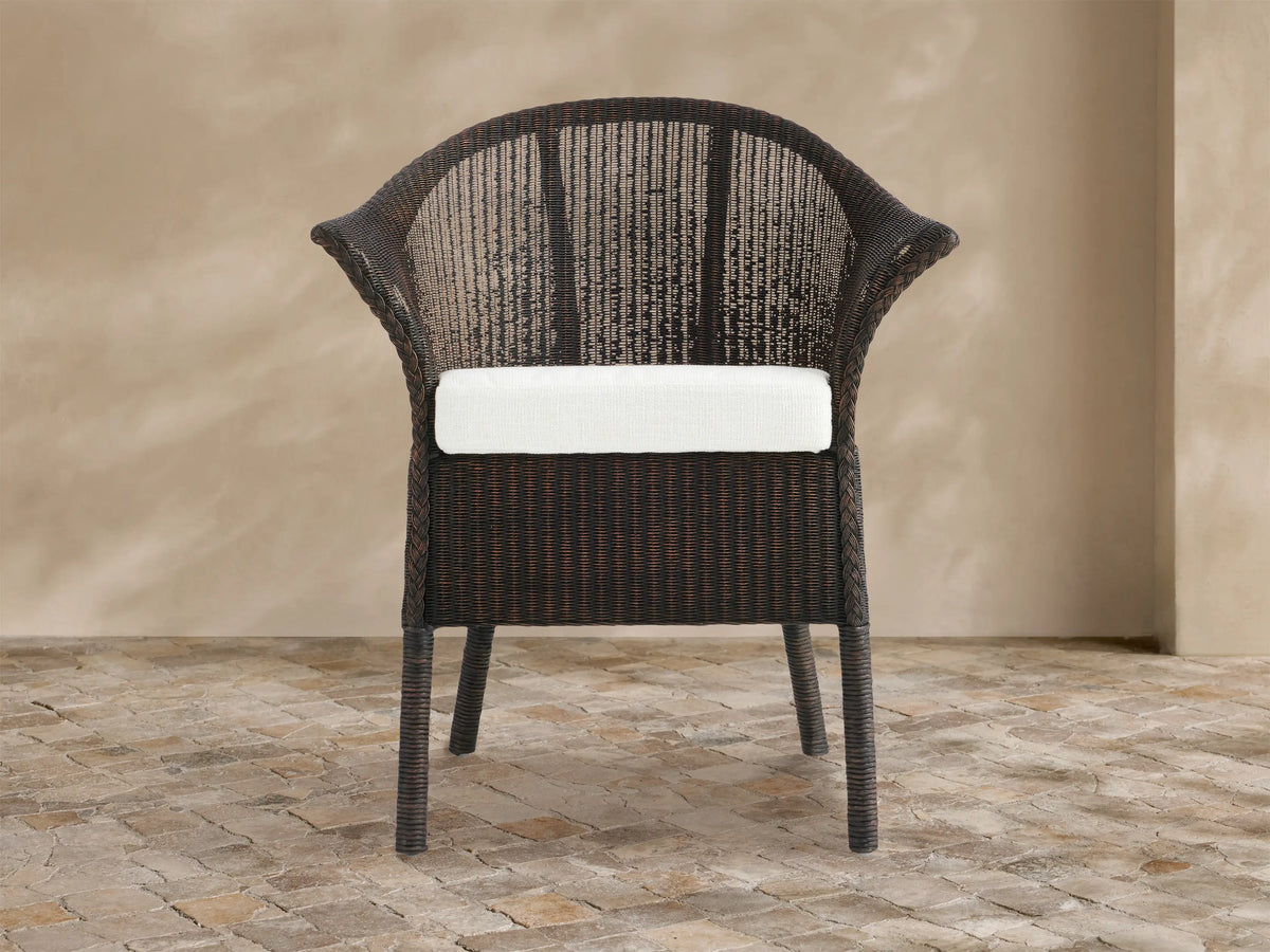 Dark brown woven rattan Nantucket Sunroom Dining Arm Chair with a white seat cushion, angled slightly on a stone floor.