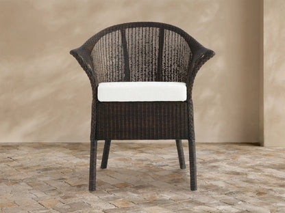 Dark brown woven rattan Nantucket Sunroom Dining Arm Chair with a white seat cushion, angled slightly on a stone floor.