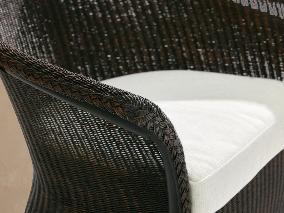 Nantucket Dining Arm Chair, dark brown woven rattan with curved back & armrests, white seat cushion, on stone floor.