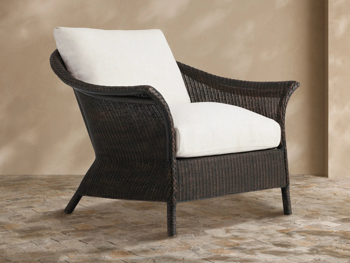 Dark brown woven Nantucket Sunroom Lounge Chair with thick white cushions, angled on a stone patio against a beige wall.