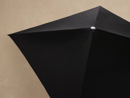 Close-up of a folded navy Nata Outdoor Square Umbrella canopy, secured by a narrow strap with a black buckle against a textured wall.
