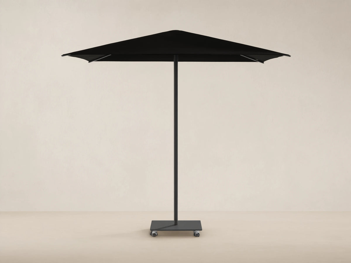 Black Nata Outdoor Square Umbrella with a square canopy, central pole, and wheeled base, against a tan wall and stone floor.
