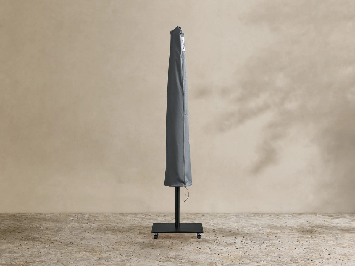 Nata Outdoor Square Scalloped Umbrella, closed. White canopy with black trim, black pole and wheeled base on a patio.