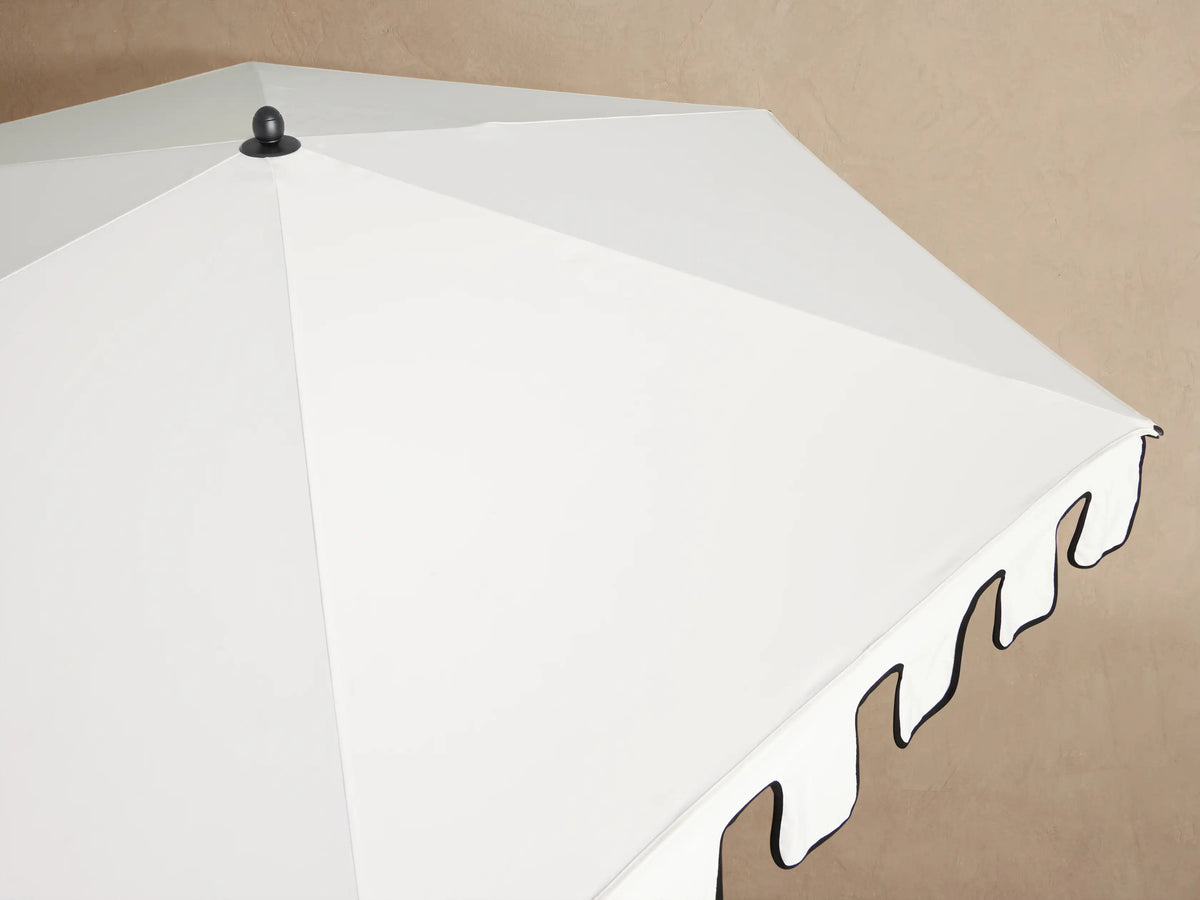 Closed Nata Round Scalloped Umbrella: white canopy with black trim, black pole & round wheeled base on patio.