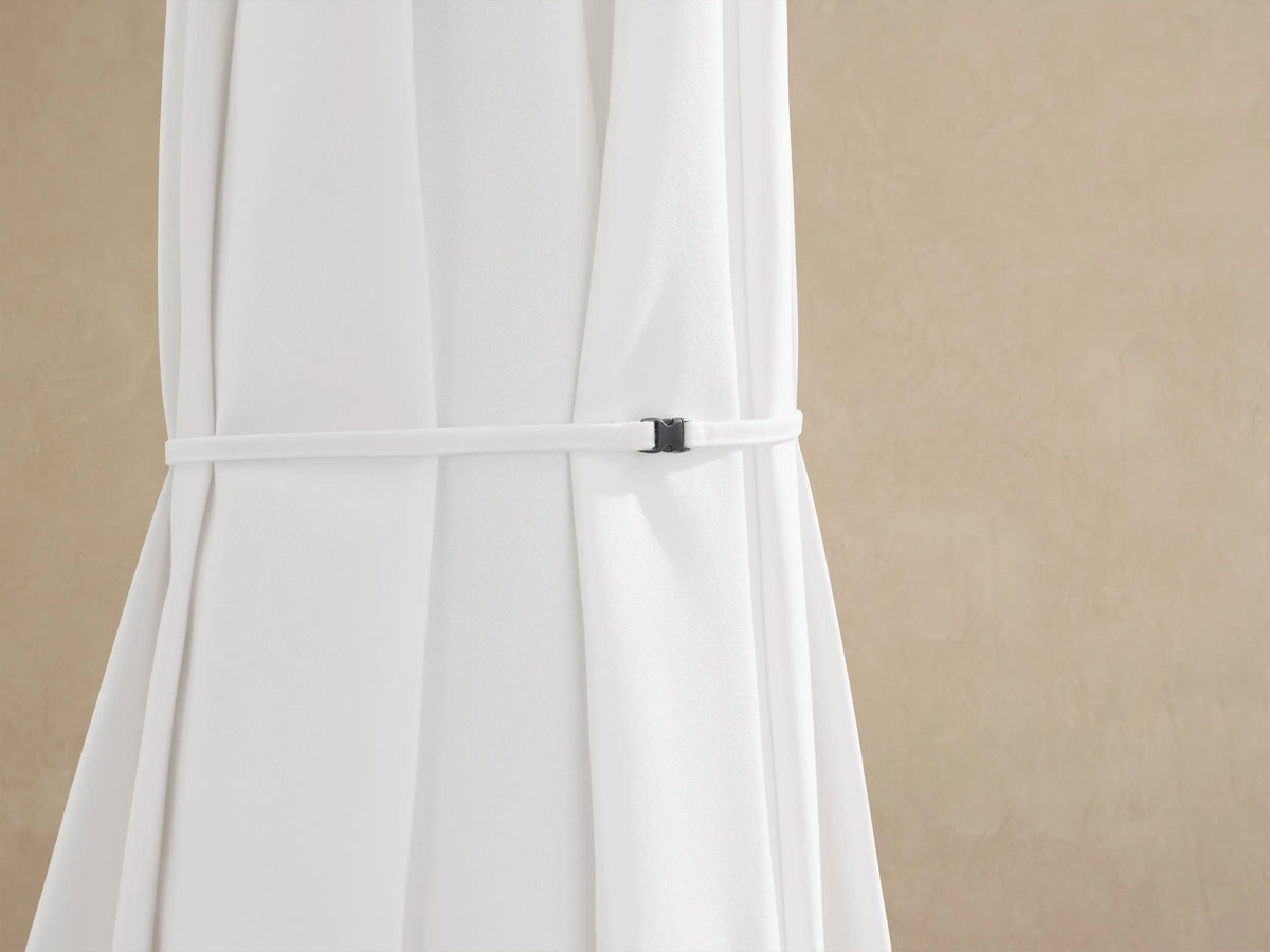 White Nata 79" Outdoor Square Umbrella, fully open, with a cream pole & wheeled base on a brick patio against a beige wall.