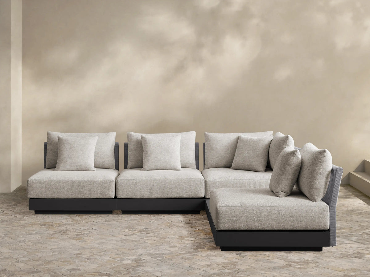 Naxos L-shaped outdoor sectional, light gray woven cushions, dark gray base, on a light tiled patio, against a beige wall.