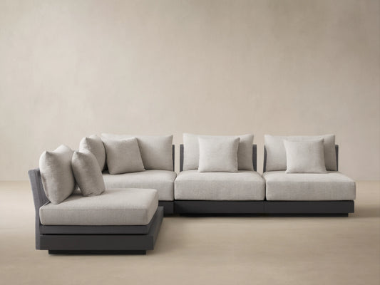 Naxos L-shaped outdoor sectional, light gray woven cushions, dark gray base, on a light tiled patio, against a beige wall.