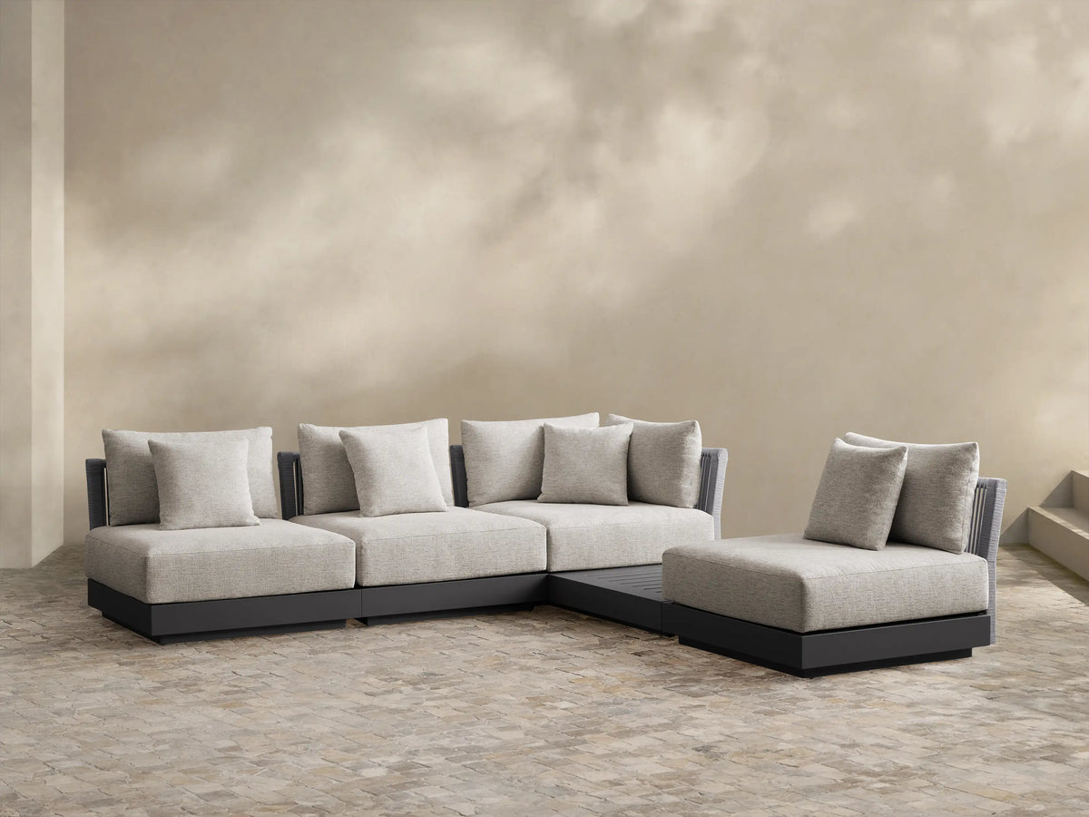 Naxos L-shaped outdoor sectional, light gray woven cushions, dark gray base, on a light tiled patio, against a beige wall.