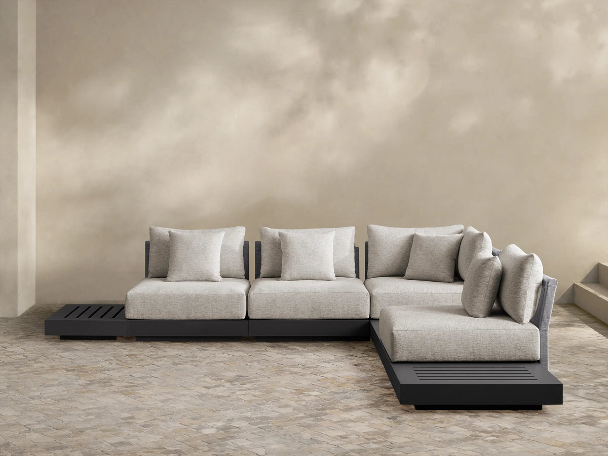Naxos Outdoor Seven-Piece Sectional: L-shaped, dark gray aluminum frame, light gray cushions, integrated slatted side table, woven armrest, on a patio.
