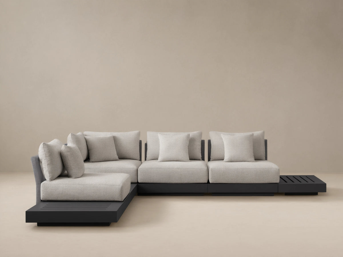Naxos Outdoor Seven-Piece Sectional: L-shaped, dark gray aluminum frame, light gray cushions, integrated slatted side table, woven armrest, on a patio.