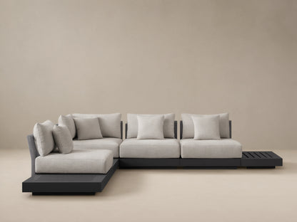 Naxos Outdoor Seven-Piece Sectional: L-shaped, dark gray aluminum frame, light gray cushions, integrated slatted side table, woven armrest, on a patio.