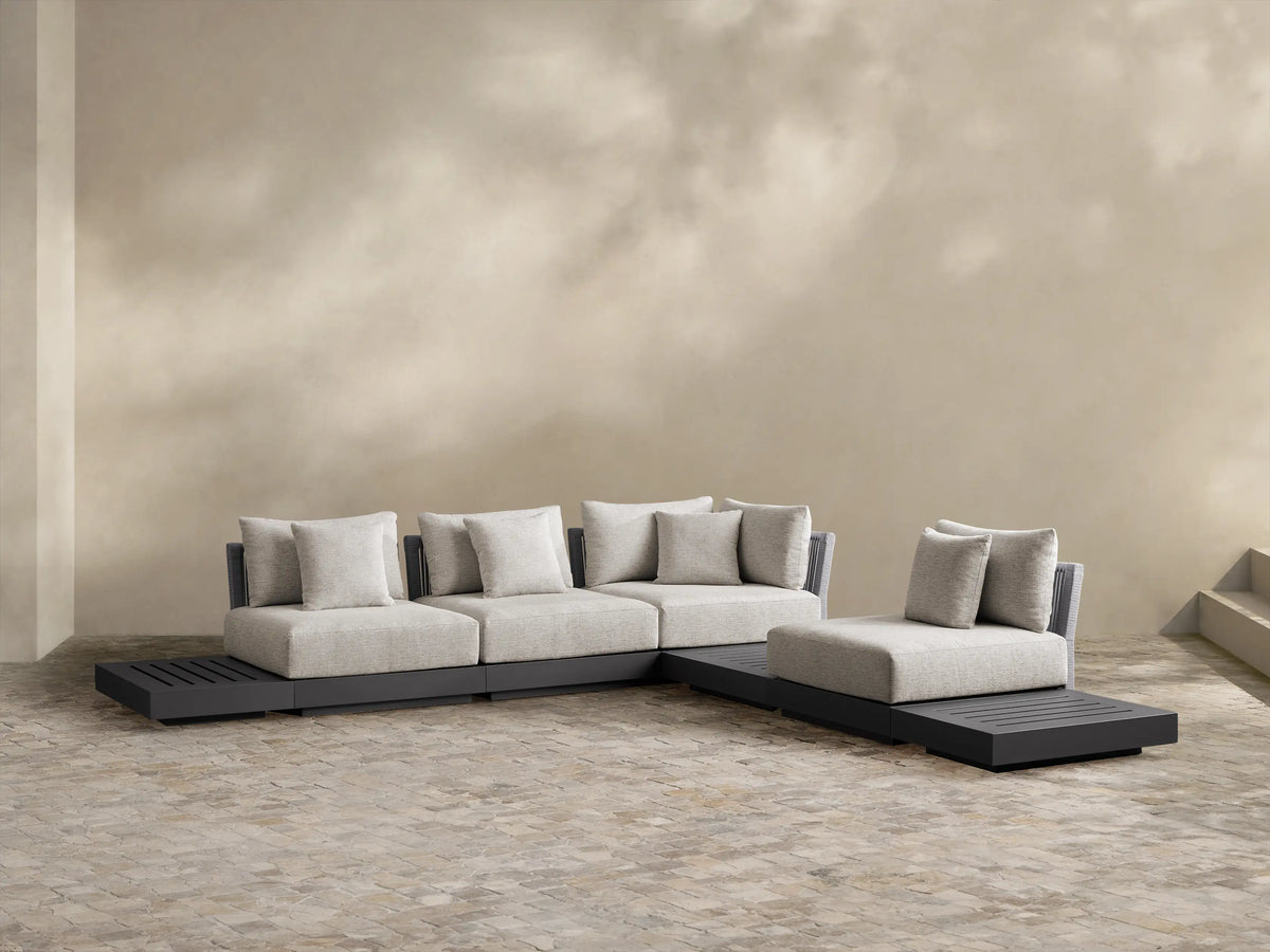 Naxos Outdoor Seven-Piece Sectional: L-shaped, dark gray aluminum frame, light gray cushions, integrated slatted side table, woven armrest, on a patio.