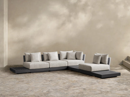 Naxos Outdoor Seven-Piece Sectional: L-shaped, dark gray aluminum frame, light gray cushions, integrated slatted side table, woven armrest, on a patio.
