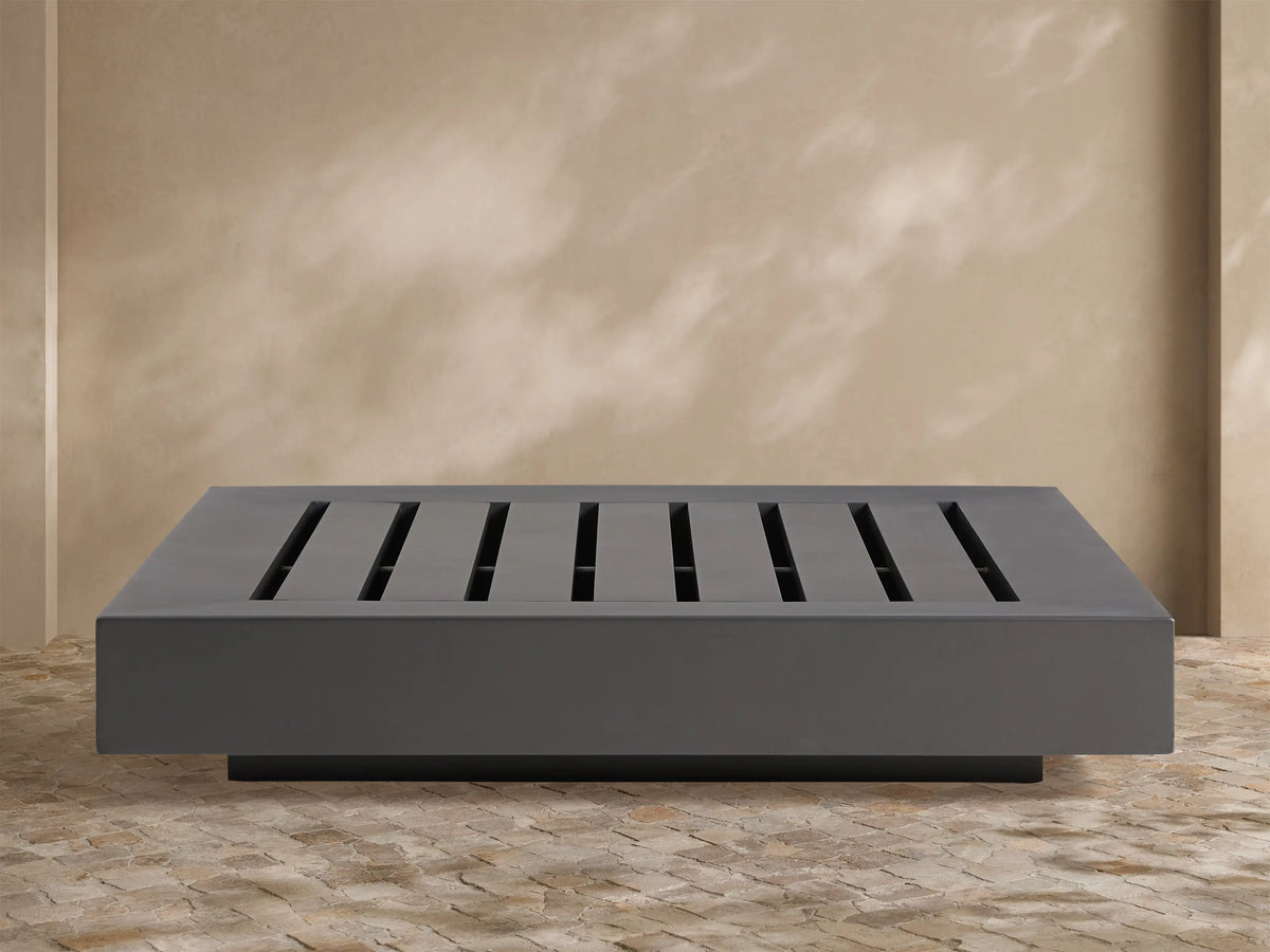 Naxos Outdoor Coffee Table: rectangular, dark gray with slatted top, on stone patio, light wall background.