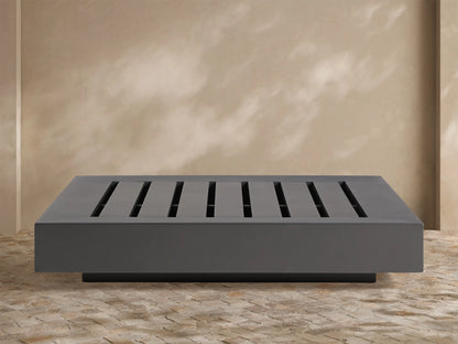 Naxos Outdoor Coffee Table: rectangular, dark gray with slatted top, on stone patio, light wall background.