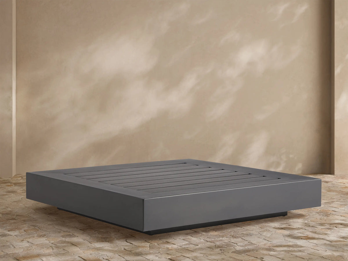 Naxos Outdoor Coffee Table: rectangular, dark gray with slatted top, on stone patio, light wall background.