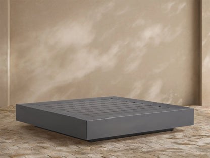 Naxos Outdoor Coffee Table: rectangular, dark gray with slatted top, on stone patio, light wall background.