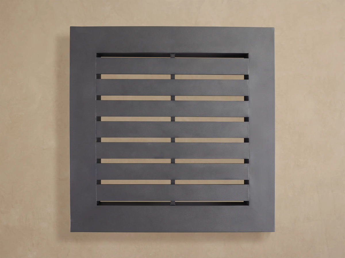Naxos Outdoor Coffee Table, dark gray square table with slatted top, sits low on a light stone patio against a beige wall.