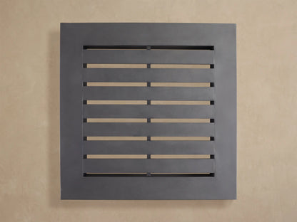 Naxos Outdoor Coffee Table, dark gray square table with slatted top, sits low on a light stone patio against a beige wall.