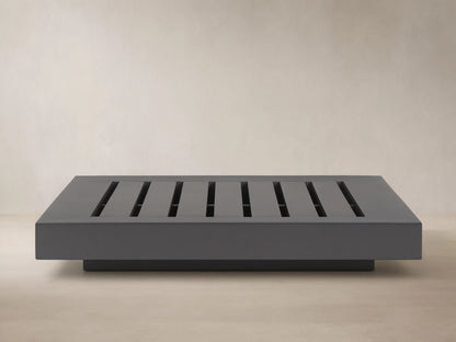 Naxos Outdoor Coffee Table: rectangular, dark gray with slatted top, on stone patio, light wall background.