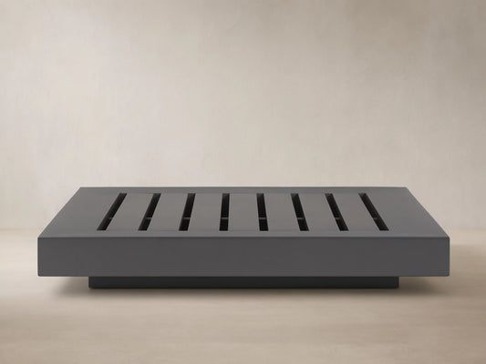 Naxos Outdoor Coffee Table: rectangular, dark gray with slatted top, on stone patio, light wall background.