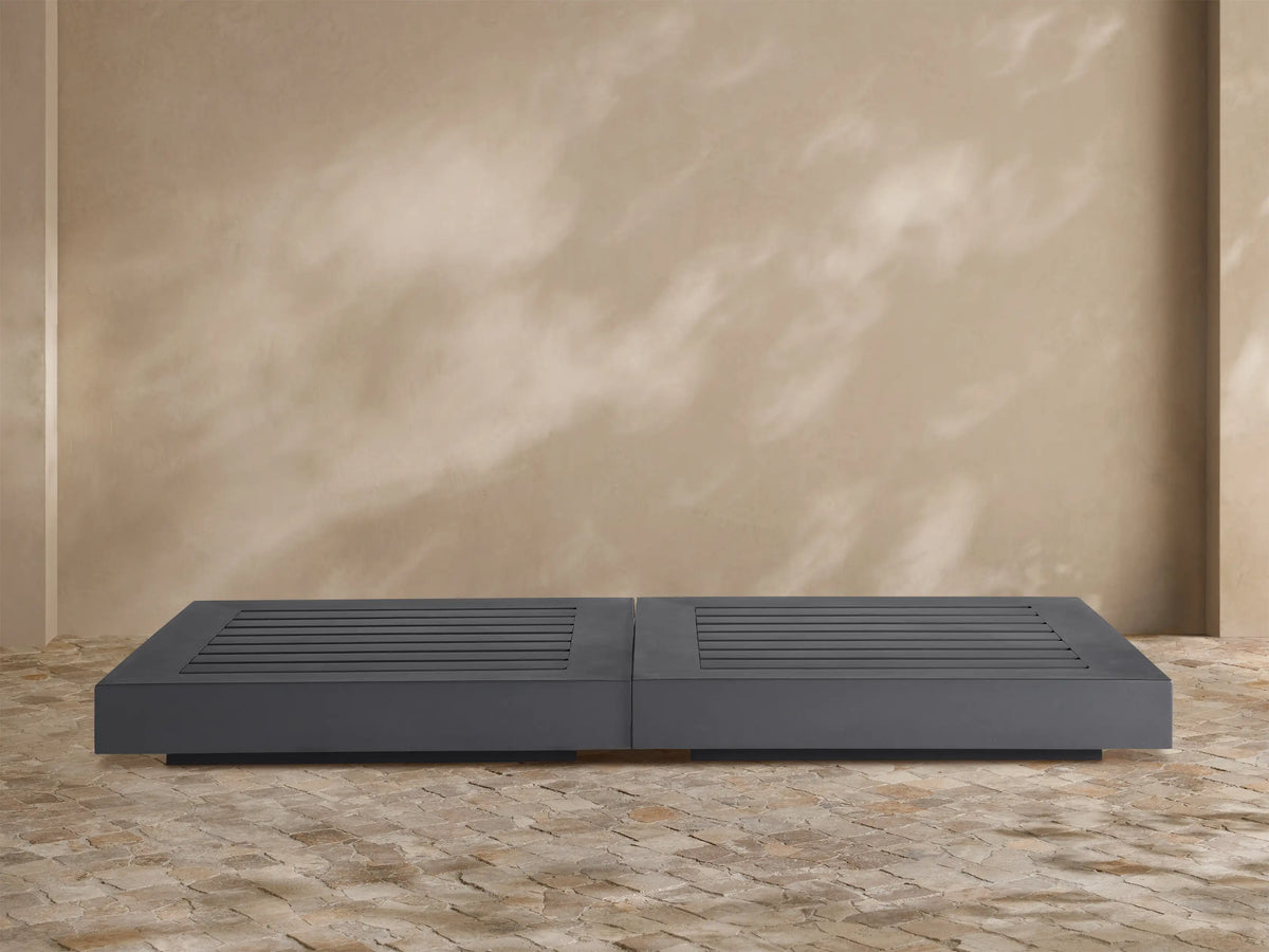 Dark gray Naxos Outdoor Double Coffee Table with slatted tops, centered on a textured stone patio against a beige wall.