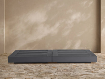 Dark gray Naxos Outdoor Double Coffee Table with slatted tops, centered on a textured stone patio against a beige wall.