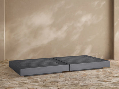 Dark gray Naxos Outdoor Double Coffee Table with slatted tops, centered on a textured stone patio against a beige wall.