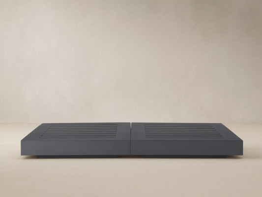 Dark gray Naxos Outdoor Double Coffee Table with slatted tops, centered on a textured stone patio against a beige wall.