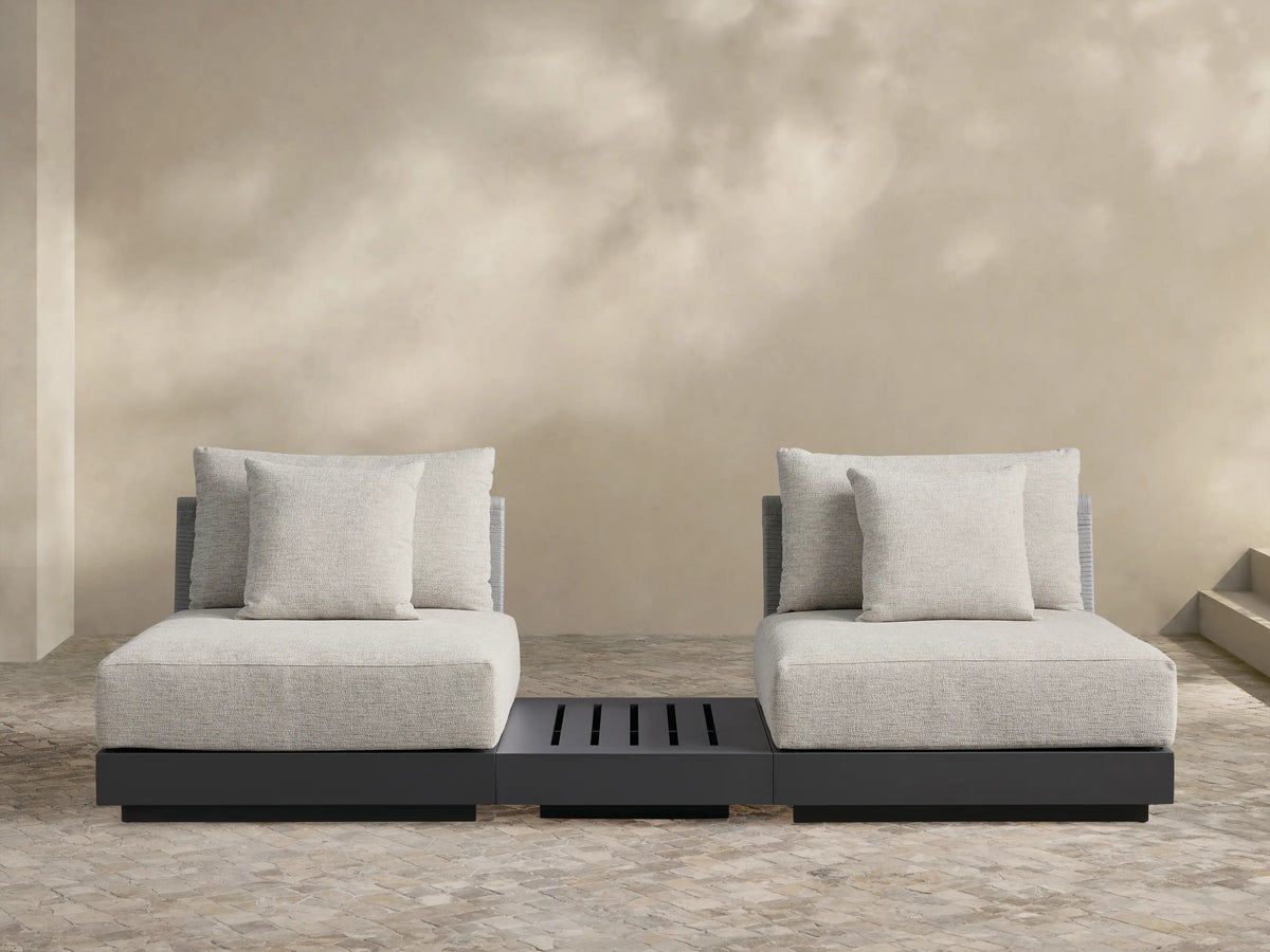 Naxos Outdoor Three-Piece Sectional with End Table. Light grey cushions on charcoal bases, centered by a slatted dark grey end table, on a stone patio.