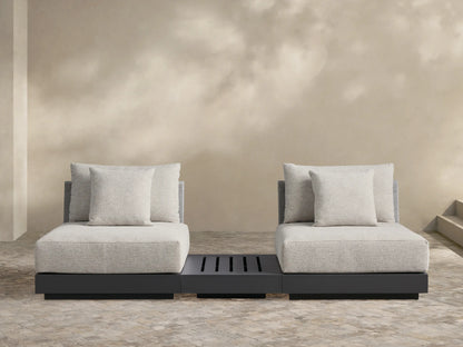 Naxos Outdoor Three-Piece Sectional with End Table. Light grey cushions on charcoal bases, centered by a slatted dark grey end table, on a stone patio.