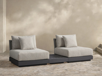 Naxos Outdoor Three-Piece Sectional with End Table. Light grey cushions on charcoal bases, centered by a slatted dark grey end table, on a stone patio.