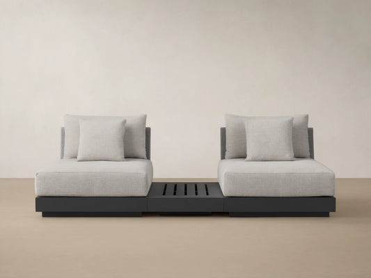 Naxos Outdoor Three-Piece Sectional with End Table. Light grey cushions on charcoal bases, centered by a slatted dark grey end table, on a stone patio.